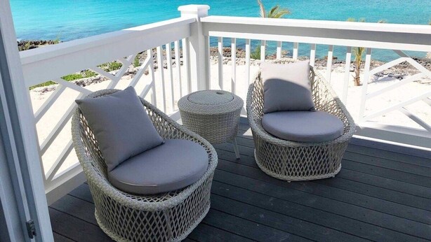 Ein Balkon mit Korbstühlen und einem kleinen runden Tisch, mit Blick auf einen Strand und klares, blaues Wasser.