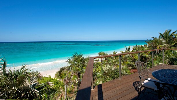 Une terrasse en bois avec une table et des chaises, donnant sur un océan turquoise et une plage de sable.