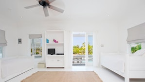 Een lichte, witte kamer met een plafondventilator, een televisie op een witte plank, en een balkon met uitzicht op palmbomen.
