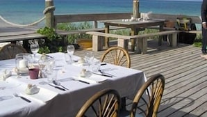 Een tafel op een houten terras met uitzicht op de oceaan, een rieten parasol en een persoon op de achtergrond.