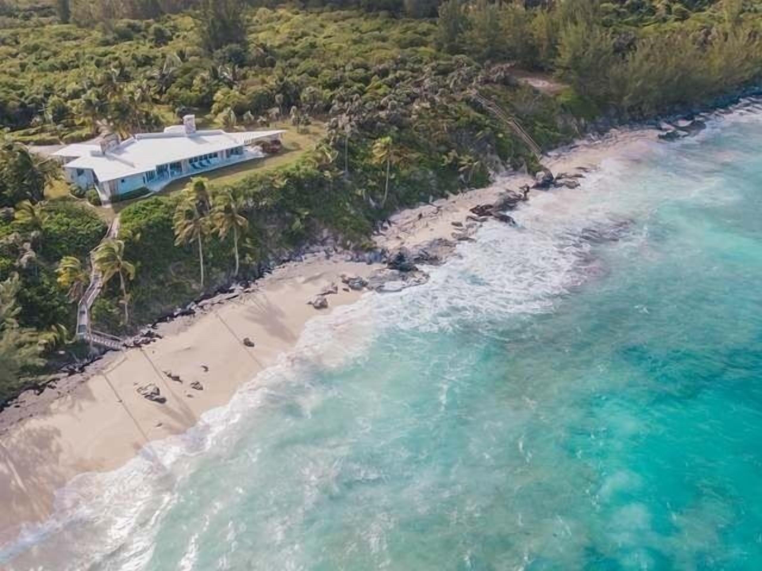 An aerial view of a beachfront property with a large house, a sandy beach, and clear turquoise waters.