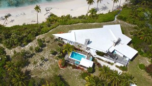 Vue aérienne d’une maison de plage moderne dotée d’une piscine, entourée de palmiers et bordée par une plage.