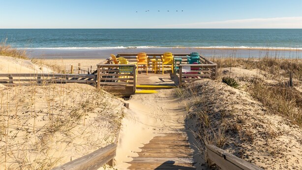 Ein hölzerner Steg führt zu einem Strandbereich mit bunten Adirondack-Stühlen und einer Holzhütte.