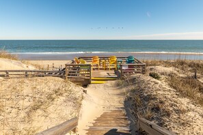 Ein Holzsteg führt zu einem Strandbereich mit bunten Adirondack-Stühlen und einem Schild, das Feuerwerkskörper verbietet.