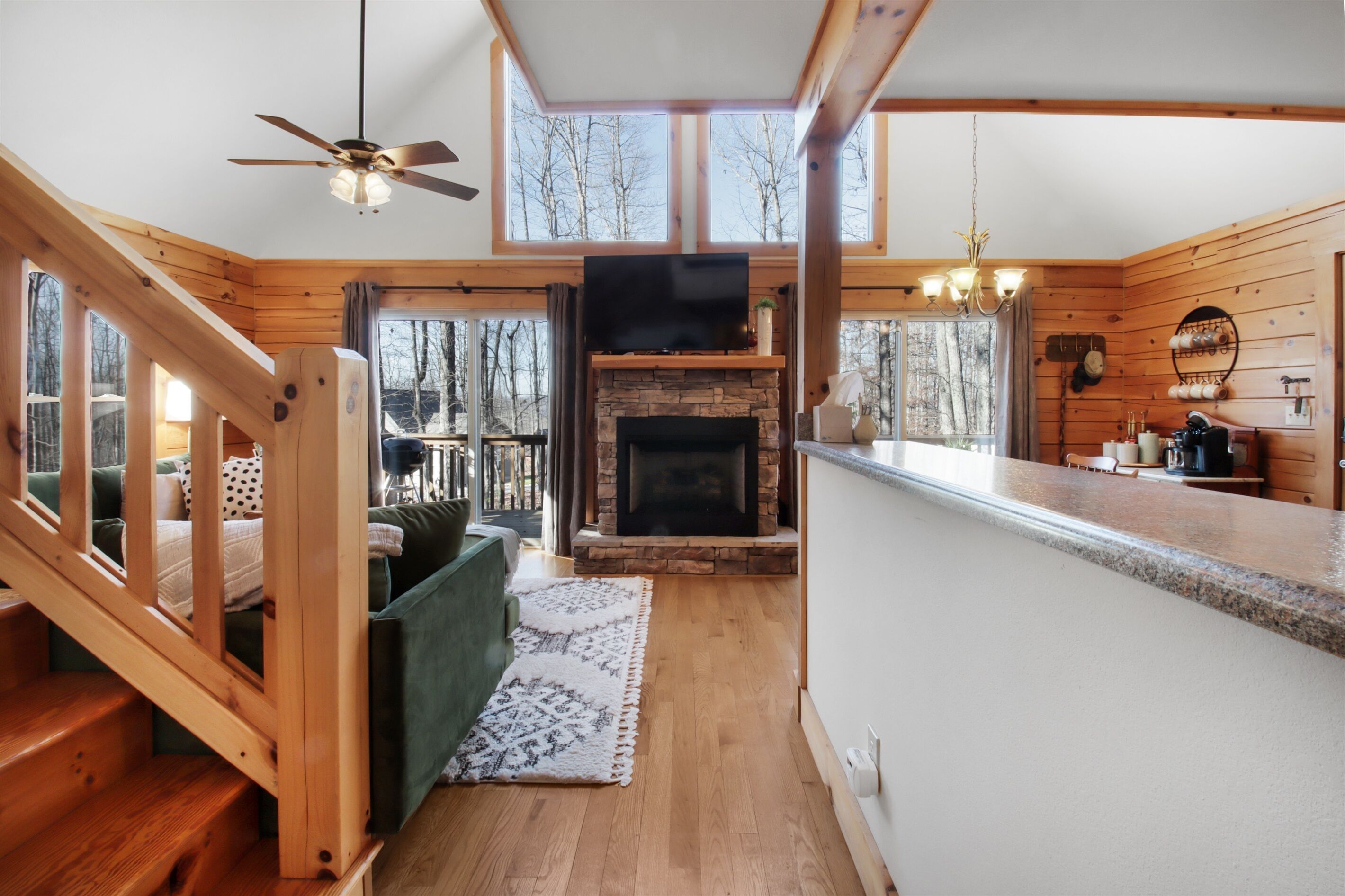 A cabin interior with a stone fireplace, wooden walls, a kitchen area, and a staircase.