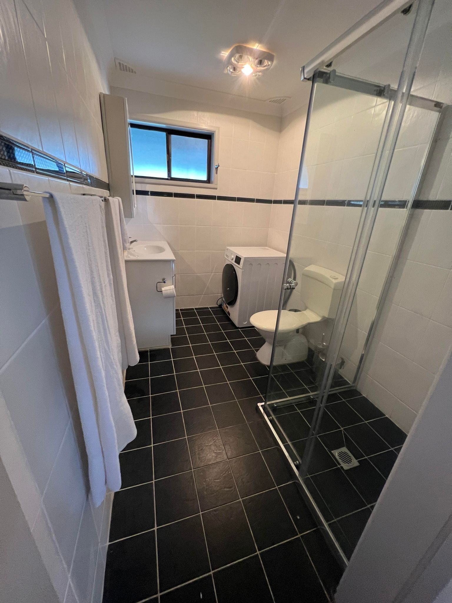 Une petite salle de bain comprenant des toilettes, un lave-linge et une douche fermée par une paroi de verre.