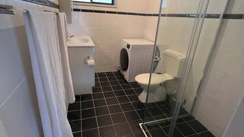 Une petite salle de bain comprenant des toilettes, un lave-linge et une douche fermée par une paroi de verre.