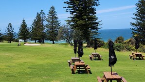 Campo de golf con mesas de picnic y sombrillas, rodeado de árboles y vistas al océano.