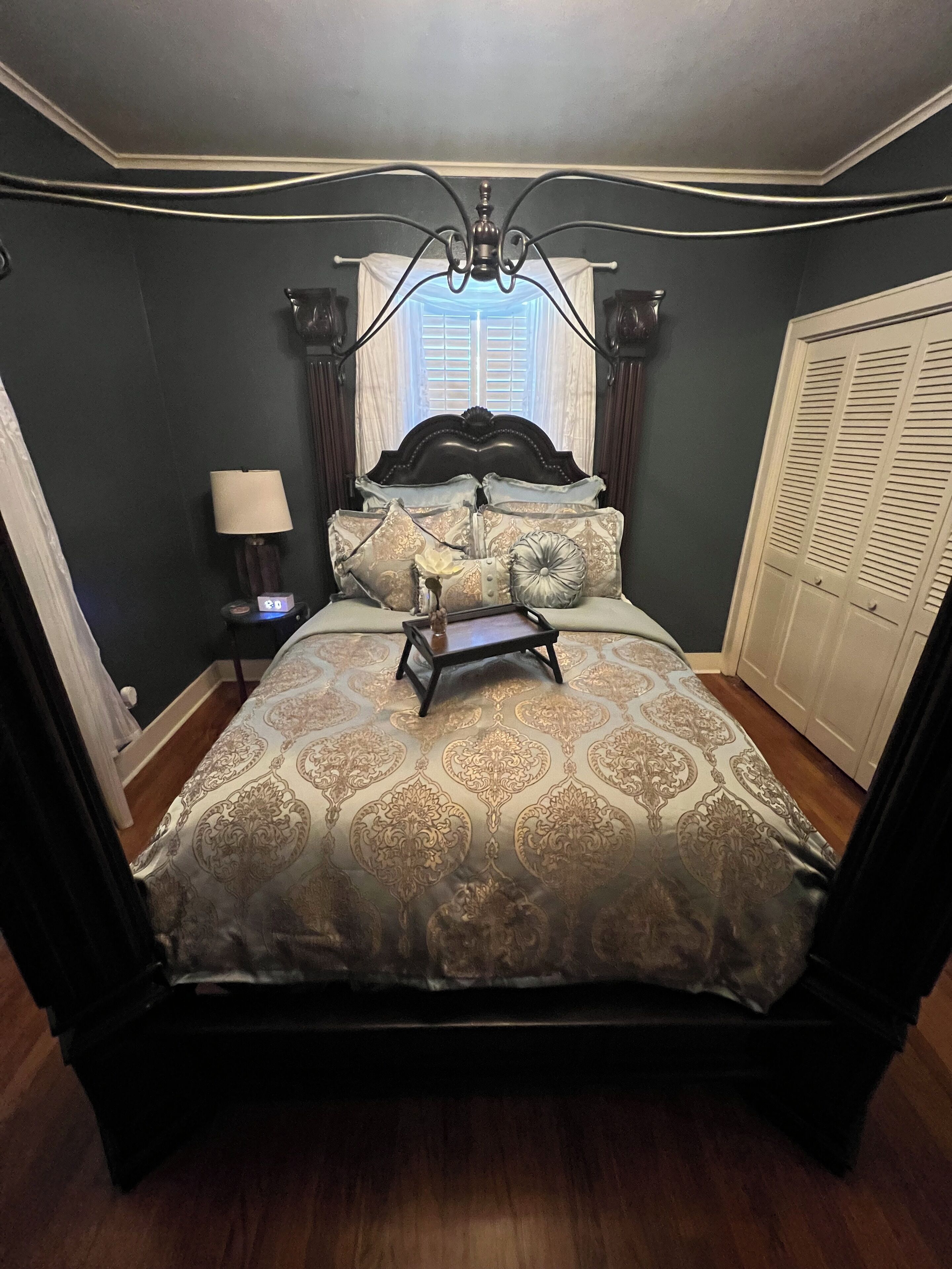 A four-poster bed with a canopy, a bedside table with a lamp, and a view of a window with curtains.