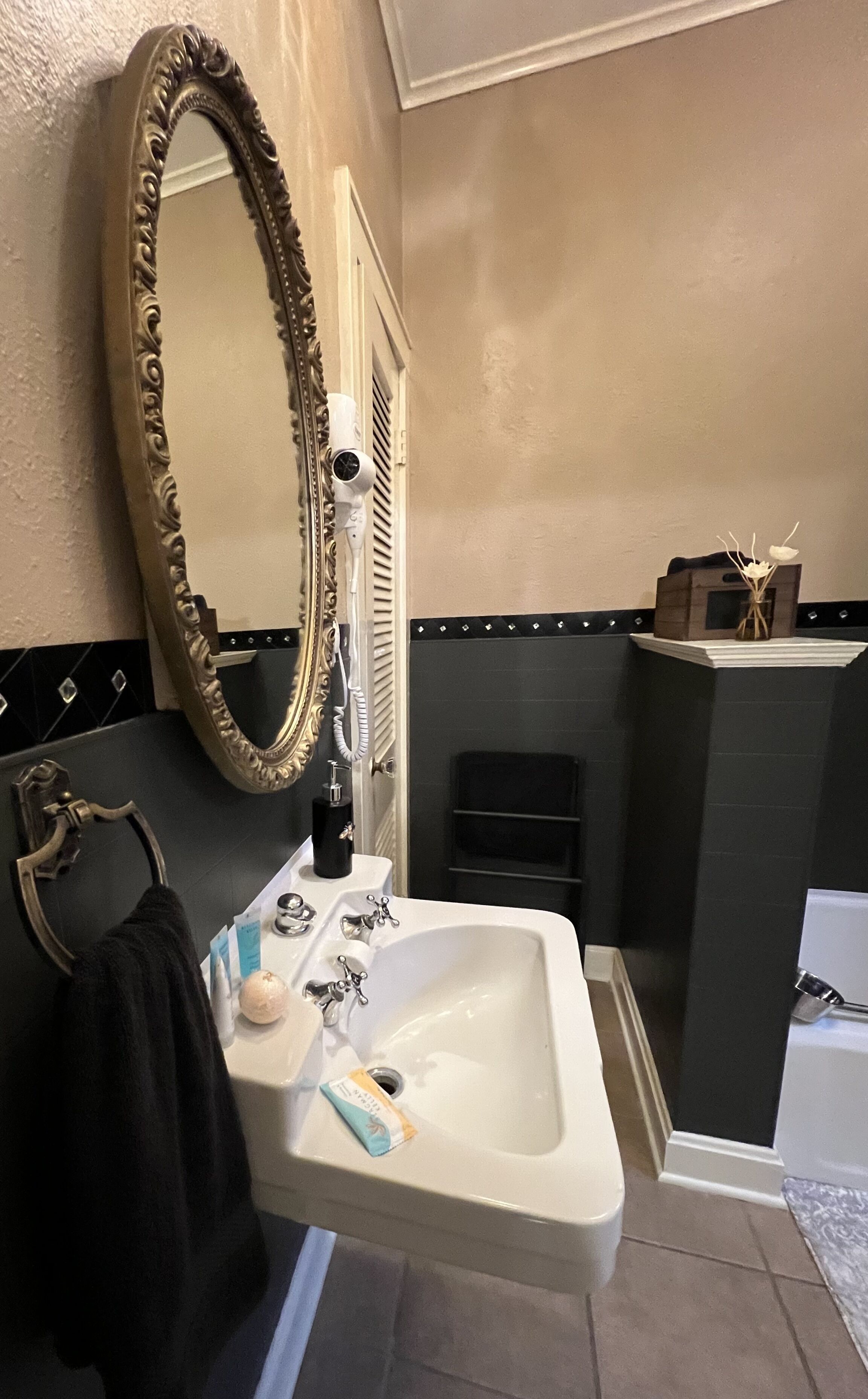 Un baño con un lavamanos blanco, un espejo grande y ornamentado, un perchero para toallas y una pequeña repisa con objetos decorativos.