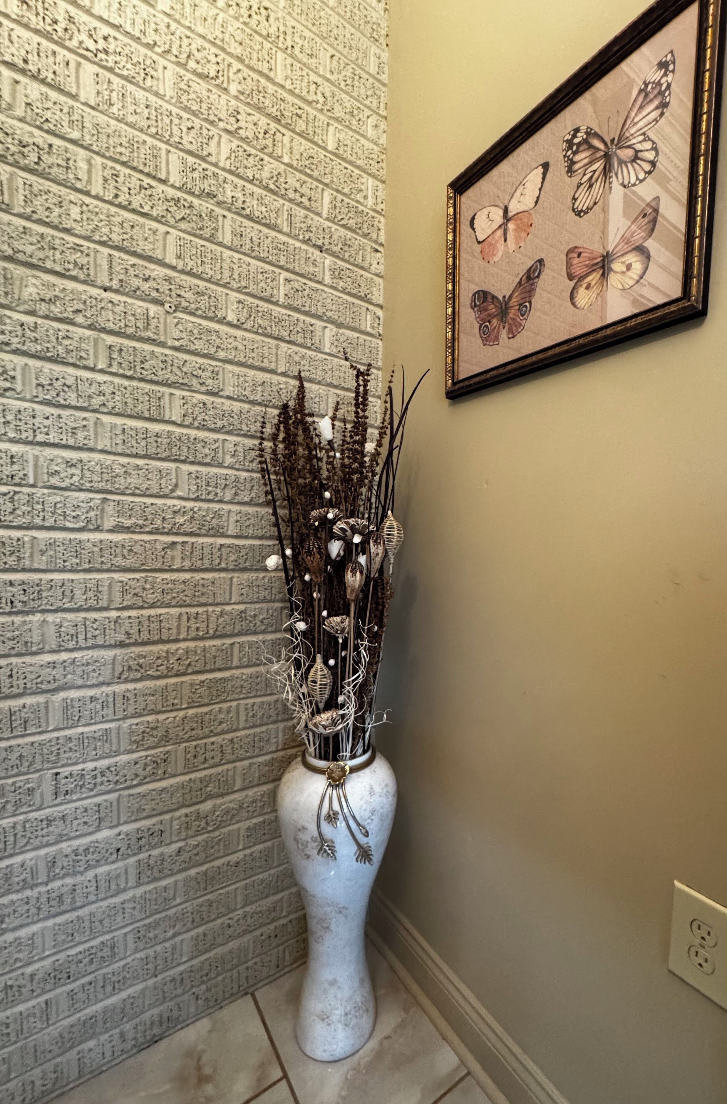 Un jarrón blanco con un arreglo floral frente a una pared de ladrillo con una obra de arte de una mariposa en marco.