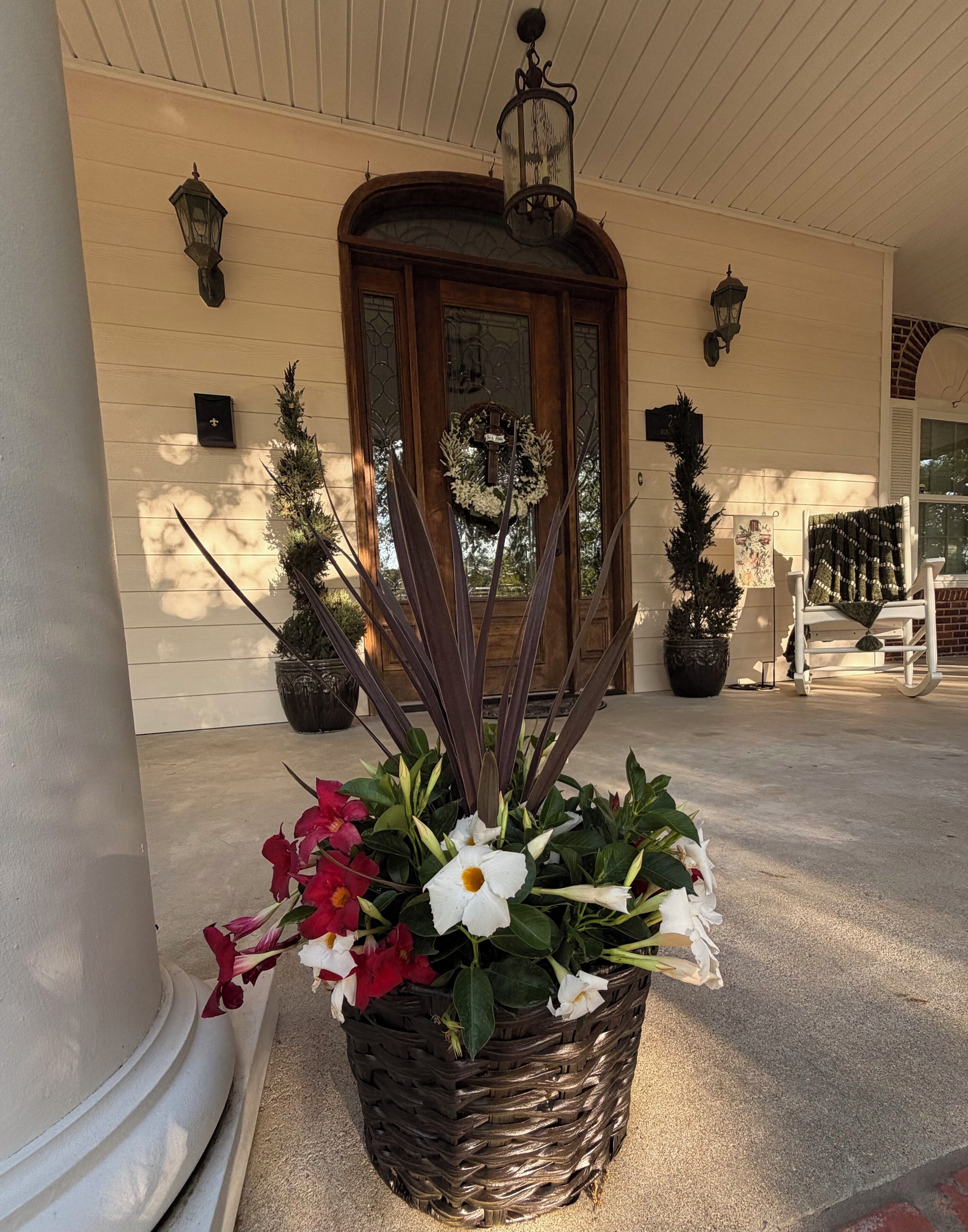 Un porche delantero con una cesta decorativa de flores, una puerta de madera con panel de vidrio y un pequeño árbol de Navidad.