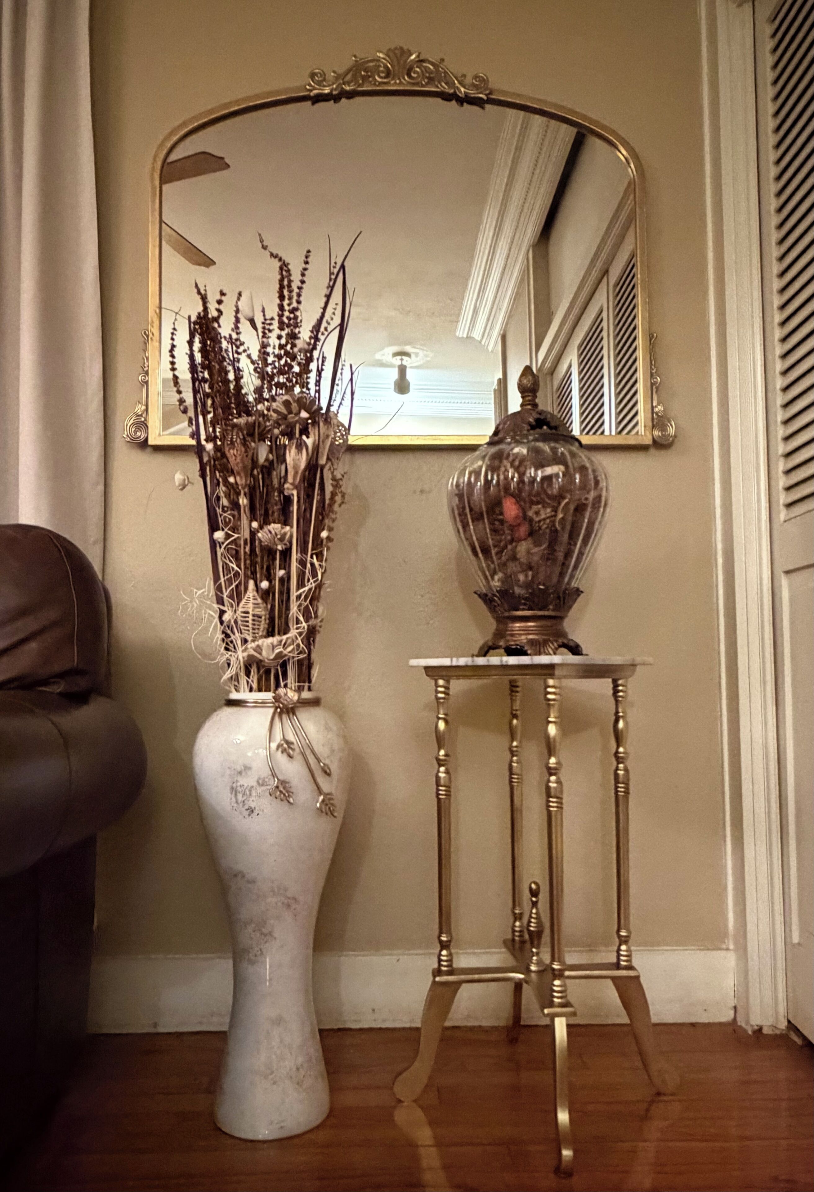A white vase with dried plants on a pedestal table next to a mirror.