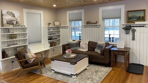 Een gezellige woonkamer met een houten plafond, witte muren en een open haard. Er zijn twee banken, een salontafel en een boekenkast.