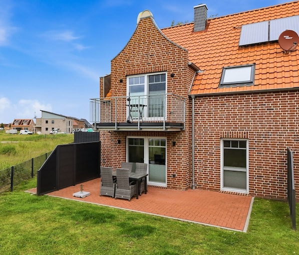 Une maison à deux étages, construite en briques, dotée d’un balcon, d’une antenne parabolique et d’une terrasse dallée aménagée avec du mobilier d’extérieur.