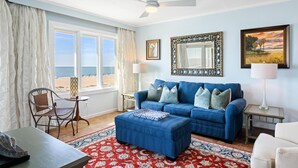 A living room with a blue sofa, a patterned rug, and a view of the beach through large windows.