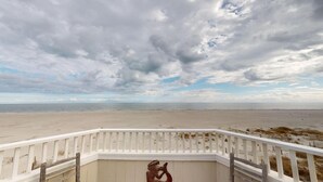 A beach view from a white balcony with a railing, overlooking a sandy beach and the ocean under a cloudy sky.