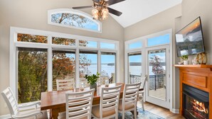 Un área de comedor con una mesa de madera y sillas blancas, ventanas amplias con vistas a árboles y una chimenea.