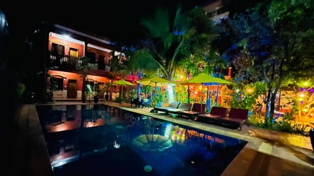 Un espace piscine nocturne avec des chaises longues et des parasols, entouré d’une végétation luxuriante et d’un bâtiment dont les fenêtres sont illuminées.