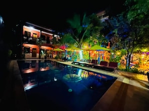 A nighttime pool area with lounge chairs and umbrellas, surrounded by lush greenery and a building with illuminated windows. - Siemreap Nitnit Boutique (Siem Reap)