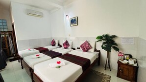 Une chambre d’hôtel avec trois lits simples, chacun doté d’un matelas blanc et agrémenté d’une décoration florale rouge. On y trouve également une armoire en bois contenant des articles de toilette et une plante en pot.