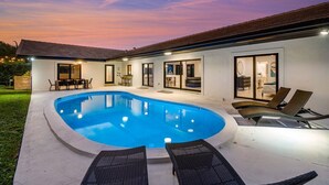 A modern house with a swimming pool, outdoor seating, and a well-lit interior.