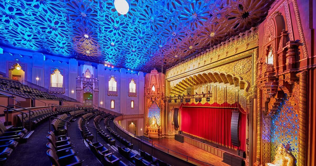 An auditorium with ornate architecture, a stage with a red curtain, and rows of seating.
