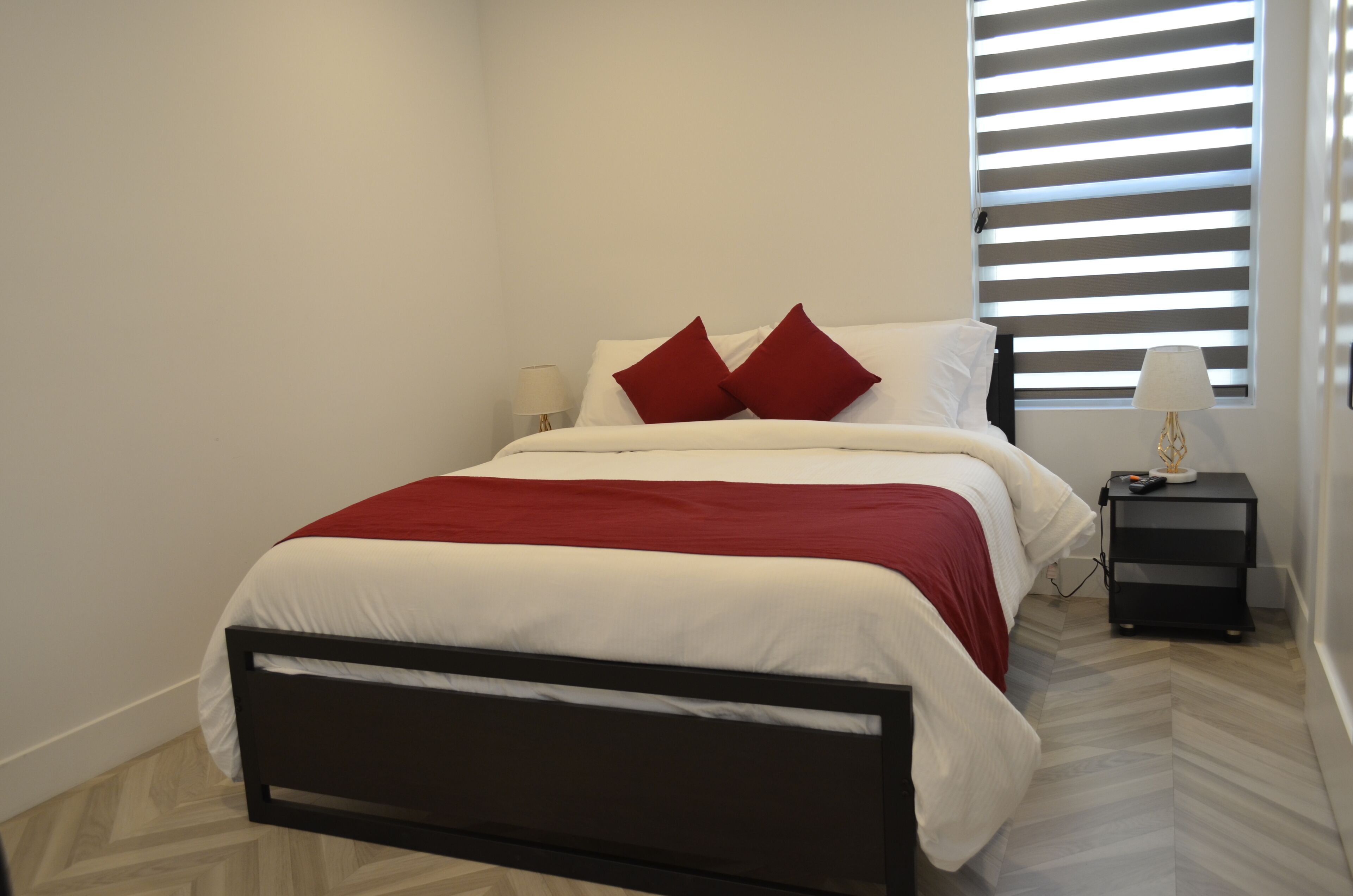 A neatly made bed with white and red bedding, a black nightstand with a lamp, and a window with blinds.