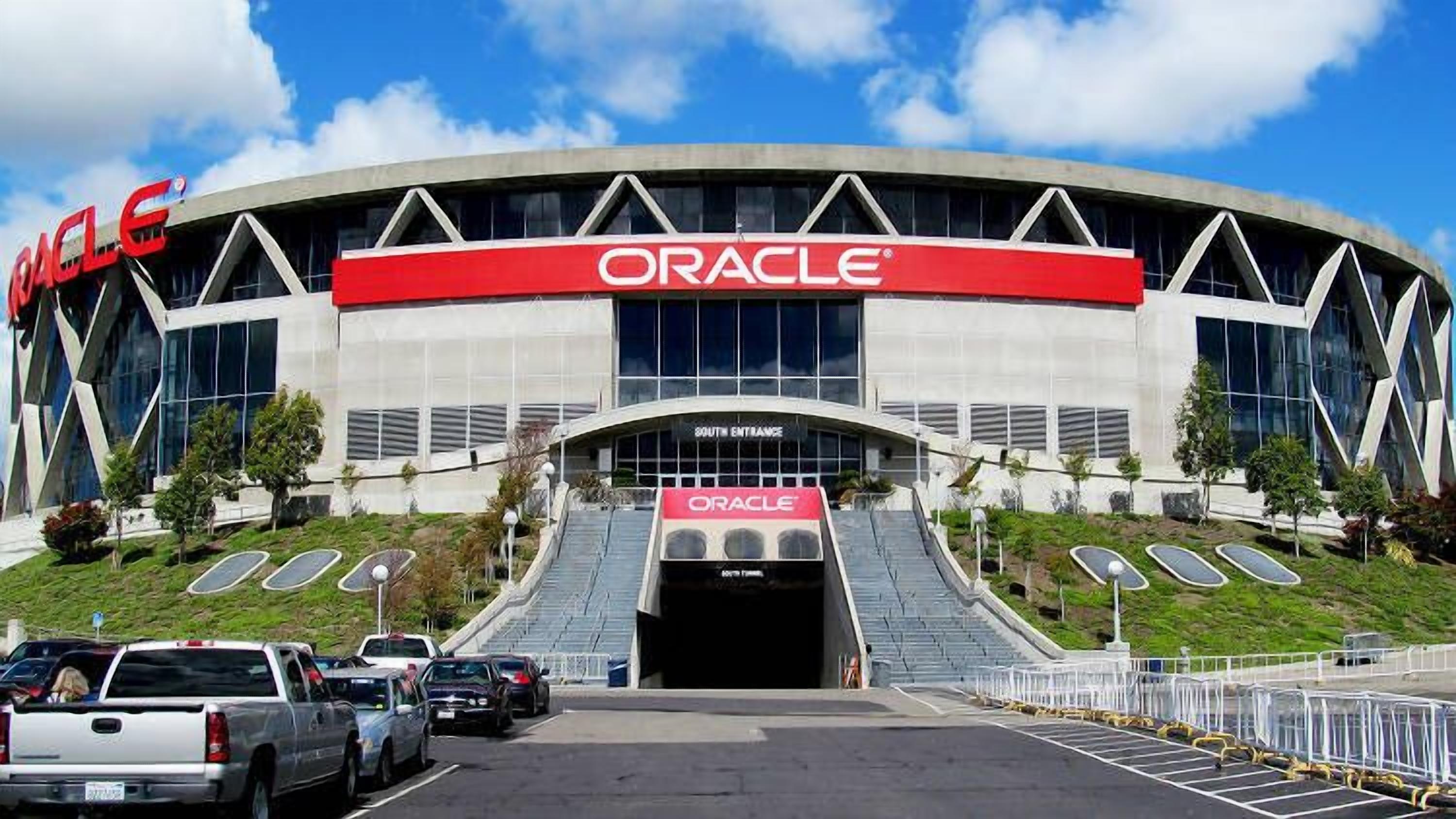 A modern building with the Oracle logo, featuring a curved entrance and a parking lot with several cars.