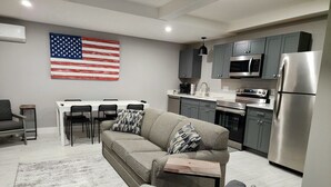 A modern living room with a gray sofa, a coffee table, and a dining area with a white table and black chairs. A kitchen with gray cabinets, a stainless steel refrigerator, and a microwave is visible. An American flag hangs on the wall. - Golf Close to lakes. 2 bedroom, 2 bath. Sleeps 6 Pool. (Mancelona)