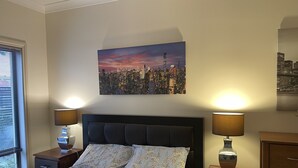 Une chambre à coucher avec un tableau représentant un paysage urbain, deux lampes de chevet, un lit avec une literie à motifs floraux et une table de chevet en bois.