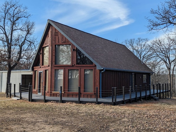 Une maison rustique en bois, dotée d’un toit à pignon abrupt, de grandes fenêtres et d’une terrasse délimitée par une clôture.