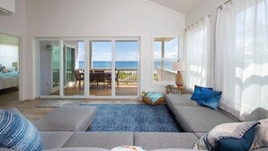 Un salón amplio con un sofá gris grande, un tapete azul y vistas al océano a través de ventanas amplias.