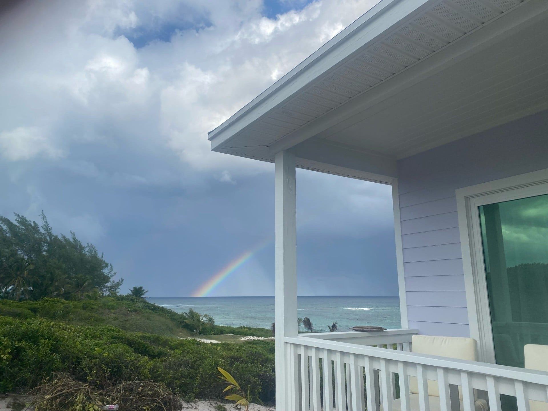 Une maison avec une véranda donnant sur une plage, un arc-en-ciel visible et une vue sur l’océan.