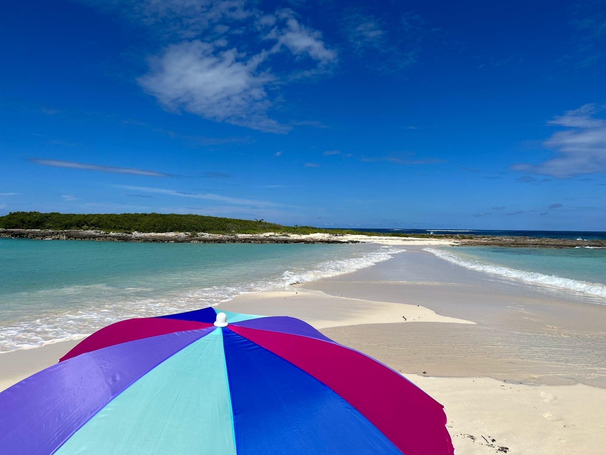 En strand med en farverig parasol i forgrunden, klar blå himmel og roligt hav.