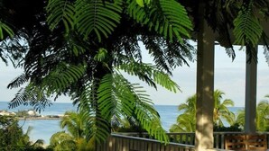 A sunlit outdoor seating area with lounge chairs and cushions, overlooking a body of water and palm trees. - Buccaneer Hill Main House 5 Bedroom Home (Governor's Harbour)