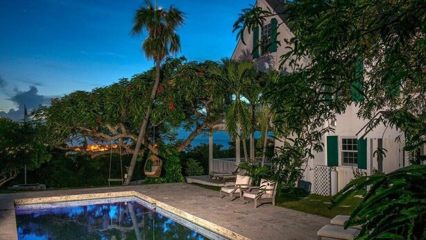 A pool area with a view of the ocean, surrounded by lush greenery and a white building with green shutters. - Buccaneer Hill Main House 5 Bedroom Home (Governor's Harbour)