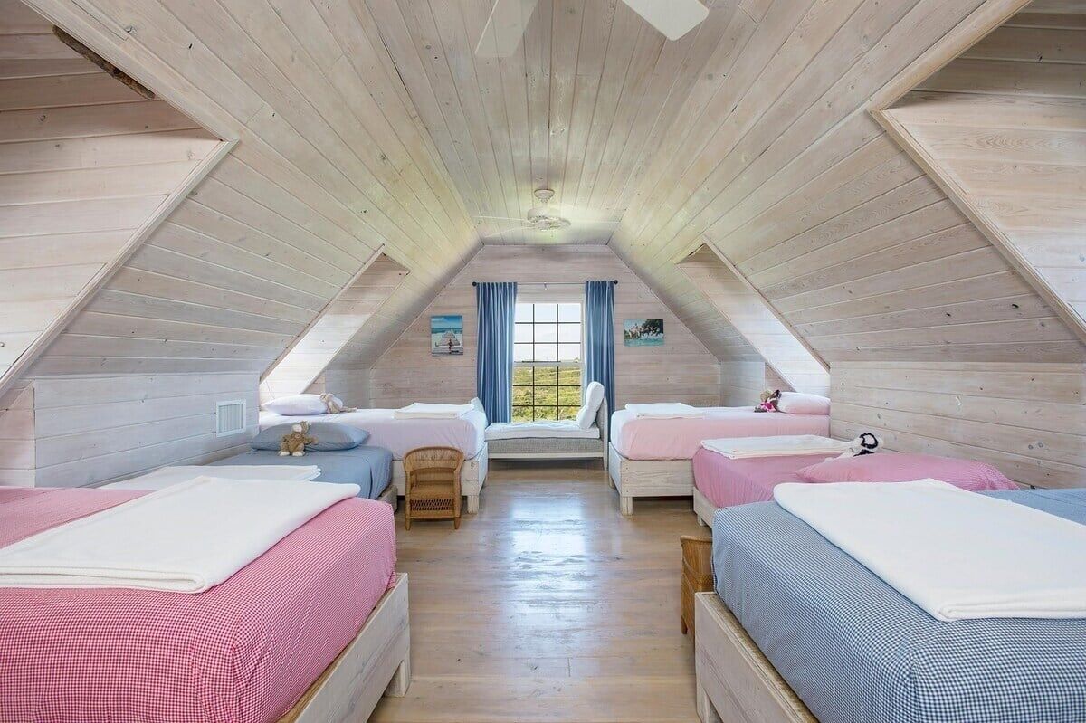 Ein gemütliches Schlafzimmer im Dachgeschoss mit Holzbalken an der Decke, zwei Betten mit rosa und blauer Bettwäsche, einem Fenster mit blauen Vorhängen und einem kleinen Stuhl.