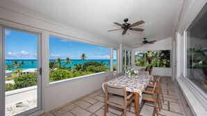 Eine überdachte Terrasse mit Tisch und Stühlen, große Fenster mit Blick auf das Meer und Palmen sowie Deckenventilatoren.