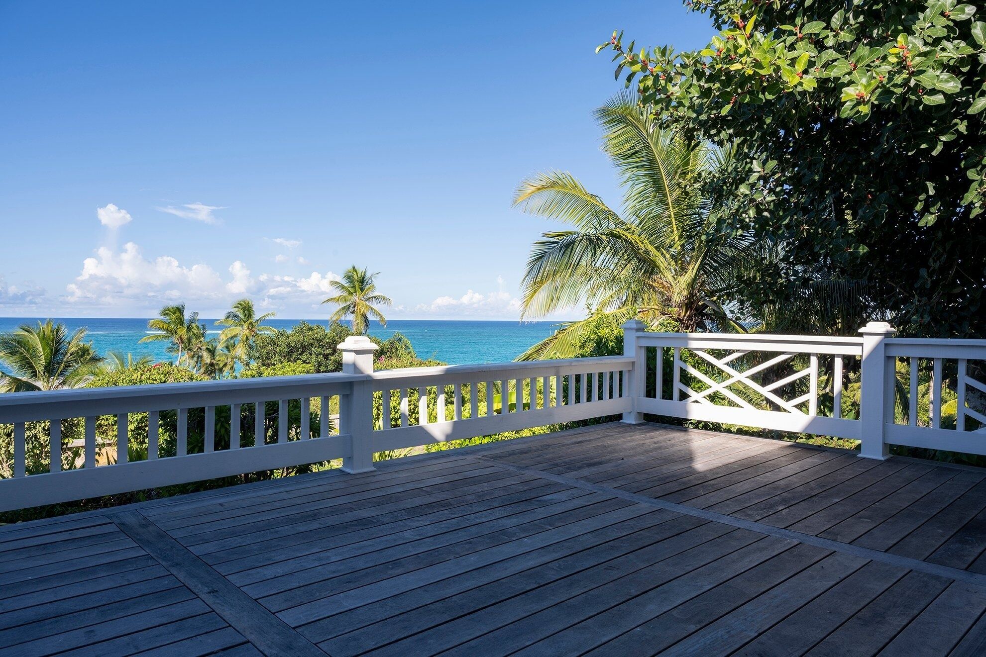 A wooden deck with a white railing overlooking a beach with palm trees and the ocean.
