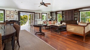 A spacious living room with a dining area, wooden flooring, and large windows offering a view of greenery.