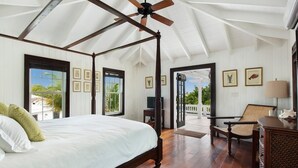 A spacious bedroom with a large bed, wooden ceiling beams, and a ceiling fan. There are framed pictures on the walls and a view of the outdoors through the windows.