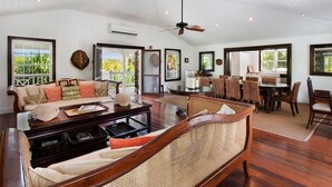 A spacious living area with a sofa, coffee table, and wicker chairs. There is a dining table with chairs, a ceiling fan, and large windows offering views of greenery.