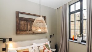 A bedroom with a bed, a hanging light fixture, a window with curtains, and a view of buildings outside. - The Old Fish Bar (Margate)