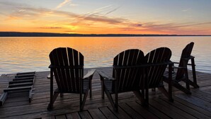Une terrasse en bois avec trois chaises Adirondack donnant sur un lac paisible au coucher du soleil.