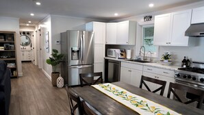 Une cuisine moderne avec des meubles blancs, des appareils en acier inoxydable et une table à manger en bois recouverte d’une nappe à motifs de feuilles de citron.