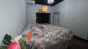Une chambre à coucher avec un lit, une fenêtre avec des stores, un luminaire suspendu et une étagère fixée au mur.