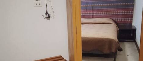 Una habitación con cama, un banco, un televisor montado en la pared y un espejo que refleja la habitación.