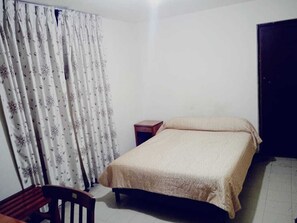Un dormitorio con una cama, una silla de madera, una mesita de noche pequeña de madera y una cortina con estampado floral.