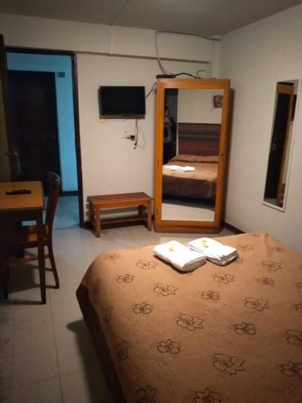 Habitación de hotel con cama, escritorio, silla, televisor y espejo.